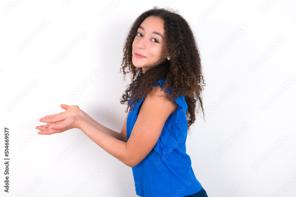 Teenager girl with afro hairstyle wearing blue T-shirt over white wall  pointing aside with hands open palms showing copy space, presenting advertisement smiling excited happy