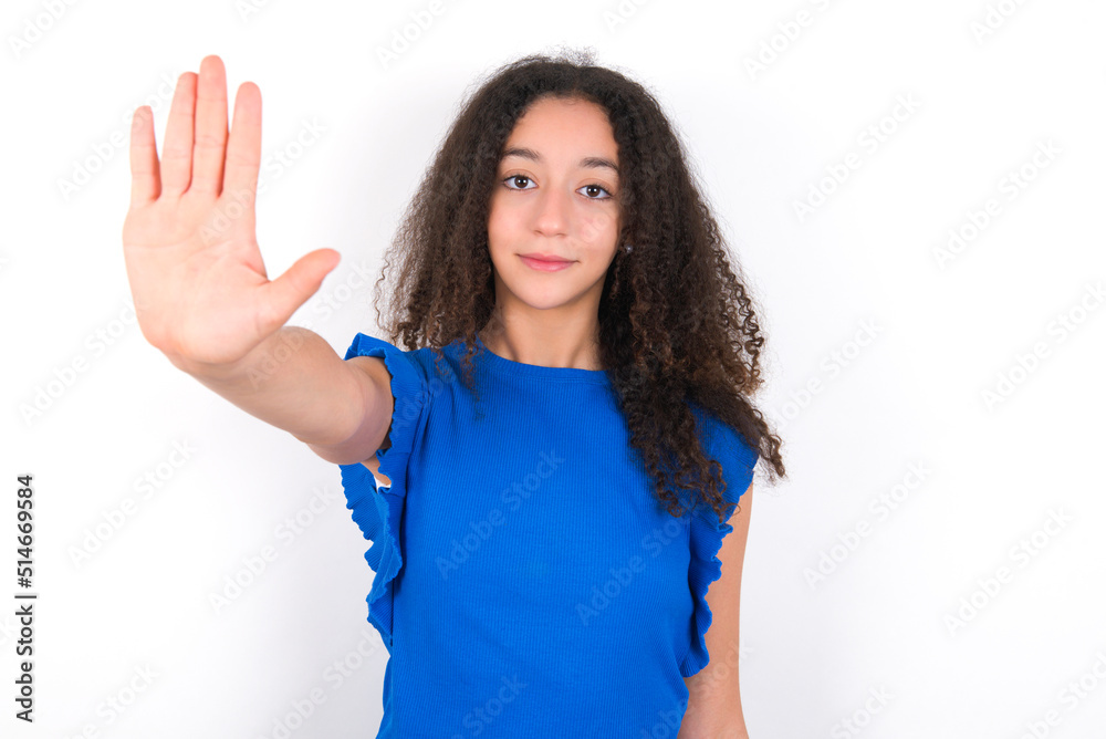 Teenager girl with afro hairstyle wearing blue T-shirt over white wall doing stop sing with palm of the hand. Warning expression with negative and serious gesture on the face. Teenager girl with afro hairstyle wearing blue T-shirt over white wall doing stop sing with palm of the hand. Warning expression with negative and serious gesture on the face.