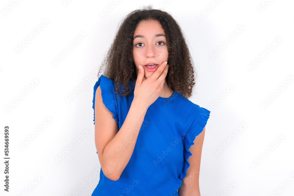 Teenager girl with afro hairstyle wearing blue T-shirt over white wall Looking fascinated with disbelief, surprise and amazed expression with hands on chin Teenager girl with afro hairstyle wearing blue T-shirt over white wall Looking fascinated with disbelief, surprise and amazed expression with hands on chin