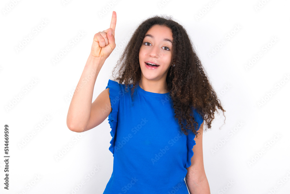 Teenager girl with afro hairstyle wearing blue T-shirt over white wall pointing finger up and looking inspired by genius thought, showing good idea sign, having clever solution in mind Teenager girl with afro hairstyle wearing blue T-shirt over white wall pointing finger up and looking inspired by genius thought, showing good idea sign, having clever solution in mind