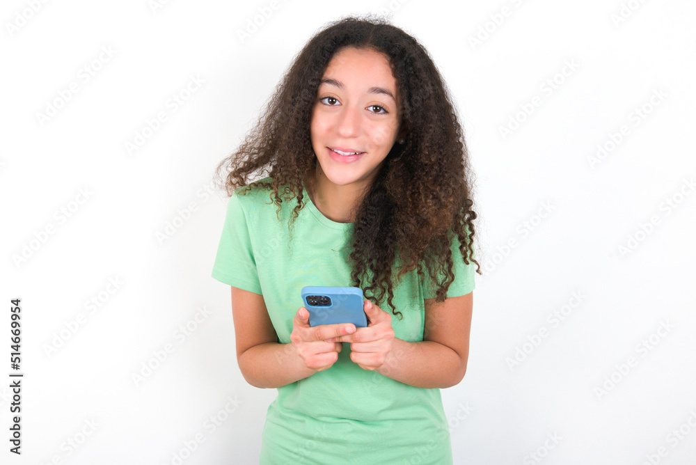 Excited Teenager girl with afro hairstyle wearing green T-shirt over white wall holding smartphone and looking amazed to the camera after receiving good news. Excited Teenager girl with afro hairstyle wearing green T-shirt over white wall holding smartphone and looking amazed to the camera after receiving good news.