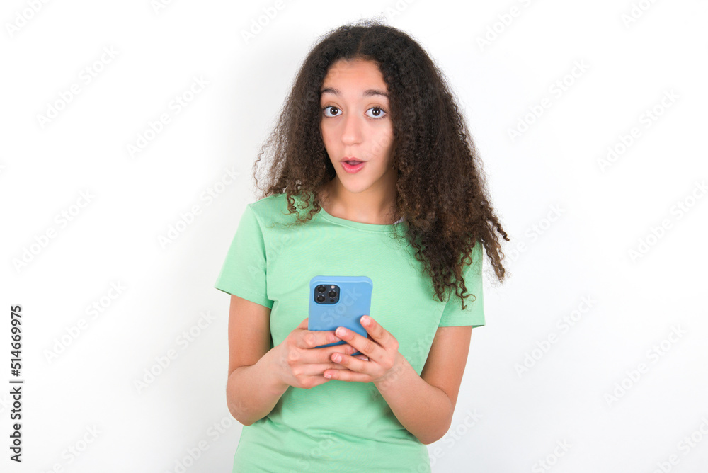 Shocked Teenager girl with afro hairstyle wearing green T-shirt over white wall opens mouth hold phone reading advert unbelievable big shopping prices Shocked Teenager girl with afro hairstyle wearing green T-shirt over white wall opens mouth hold phone reading advert unbelievable big shopping prices
