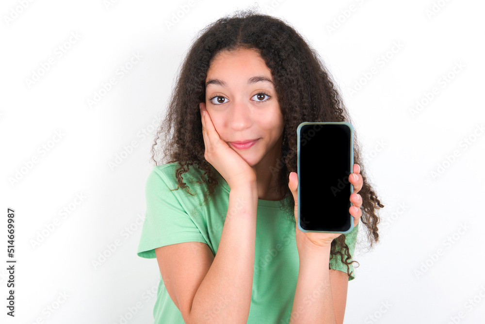 Teenager girl with afro hairstyle wearing green T-shirt over white wall hold hand modern technology use touch face palm astonished impressed scream wow omg unbelievable unexpected Teenager girl with afro hairstyle wearing green T-shirt over white wall hold hand modern technology use touch face palm astonished impressed scream wow omg unbelievable unexpected