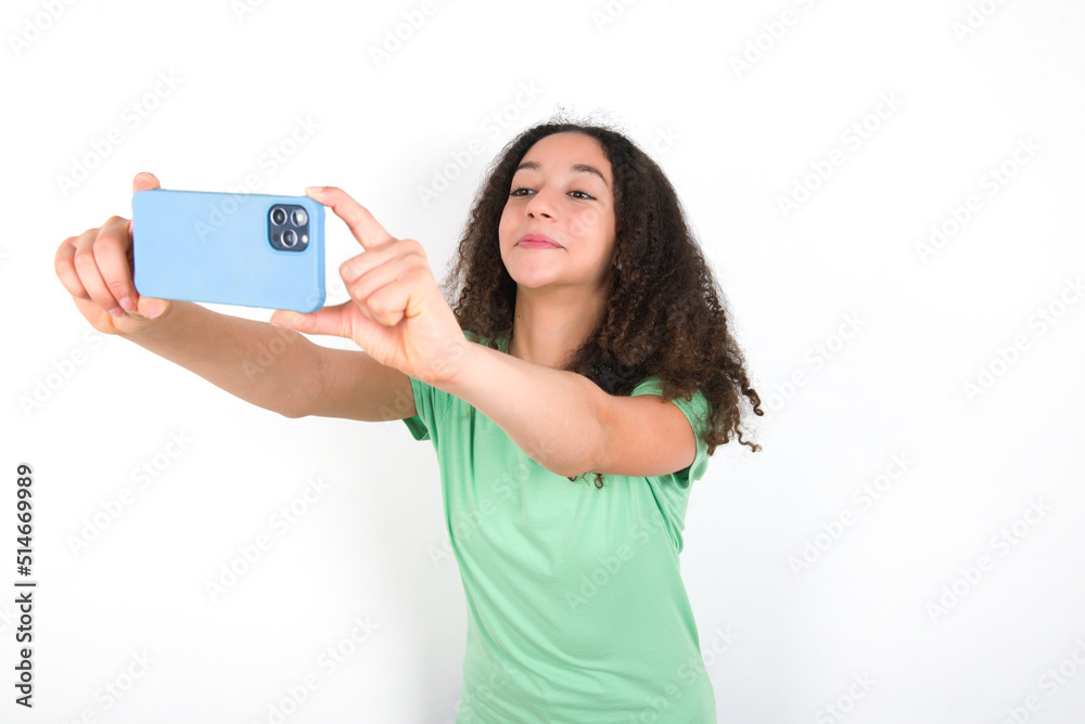 Teenager girl with afro hairstyle wearing green T-shirt over white wall taking a selfie to post it on social media or having a video call with friends. Teenager girl with afro hairstyle wearing green T-shirt over white wall taking a selfie to post it on social media or having a video call with friends.