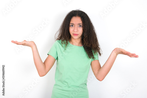 Indignant Teenager girl with afro hairstyle wearing green T-shirt over white wall gestures in bewilderment, frowns face with dissatisfaction.