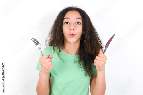 hungry Teenager girl with afro hairstyle wearing green T-shirt over white wall holding in hand fork knife want tasty yummy pizza pie