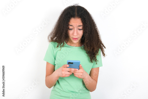 Focused Teenager girl with afro hairstyle wearing green T-shirt over white wall use smartphone reading social media news, or important e-mail