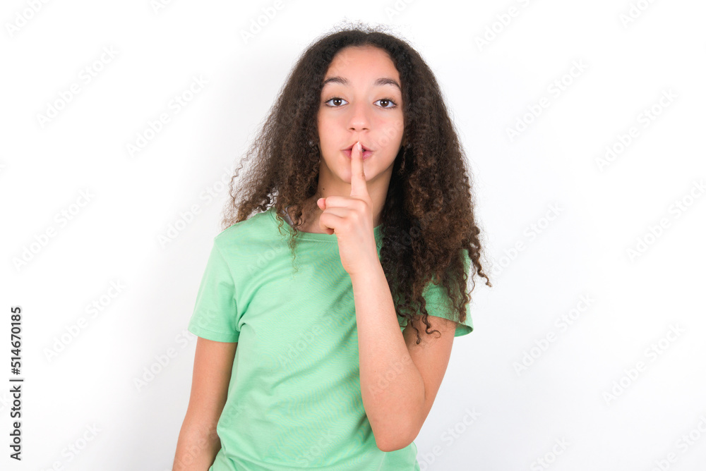 Surprised Teenager girl with afro hairstyle wearing green T-shirt over white wall makes silence gesture, keeps finger over lips and looks mysterious at camera Surprised Teenager girl with afro hairstyle wearing green T-shirt over white wall makes silence gesture, keeps finger over lips and looks mysterious at camera