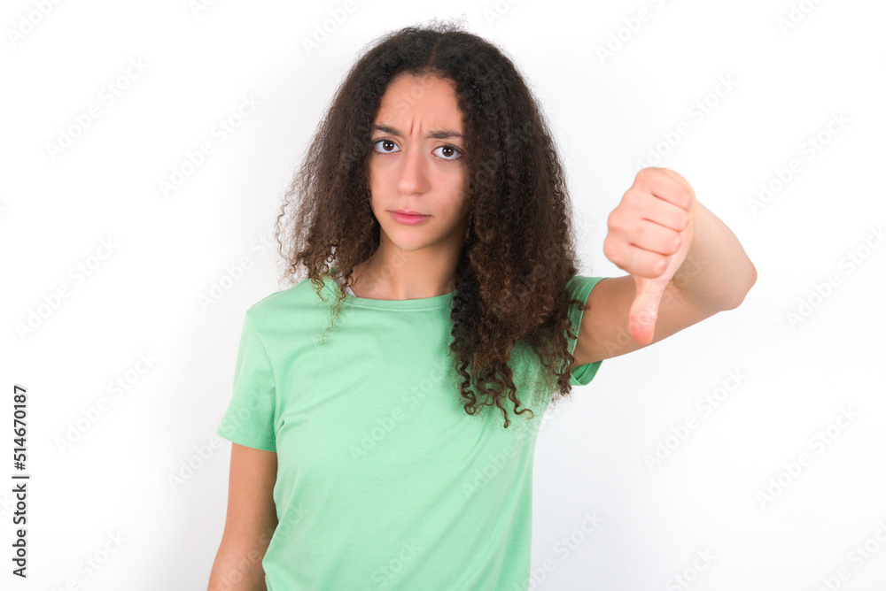 Teenager girl with afro hairstyle wearing green T-shirt over white wall looking unhappy and angry showing rejection and negative with thumbs down gesture. Bad expression. Teenager girl with afro hairstyle wearing green T-shirt over white wall looking unhappy and angry showing rejection and negative with thumbs down gesture. Bad expression.