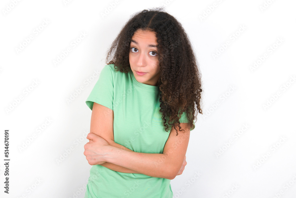 Teenager girl with afro hairstyle wearing green T-shirt over white wall shaking and freezing for winter cold with sad and shock expression on face. Teenager girl with afro hairstyle wearing green T-shirt over white wall shaking and freezing for winter cold with sad and shock expression on face.