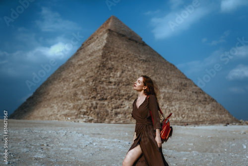 Young redhead tourist girl in brawn dress with a leather backpack standing on the sand in Egypt, Cairo - Giza. Pyramid of Khafre on backround. Copy space
