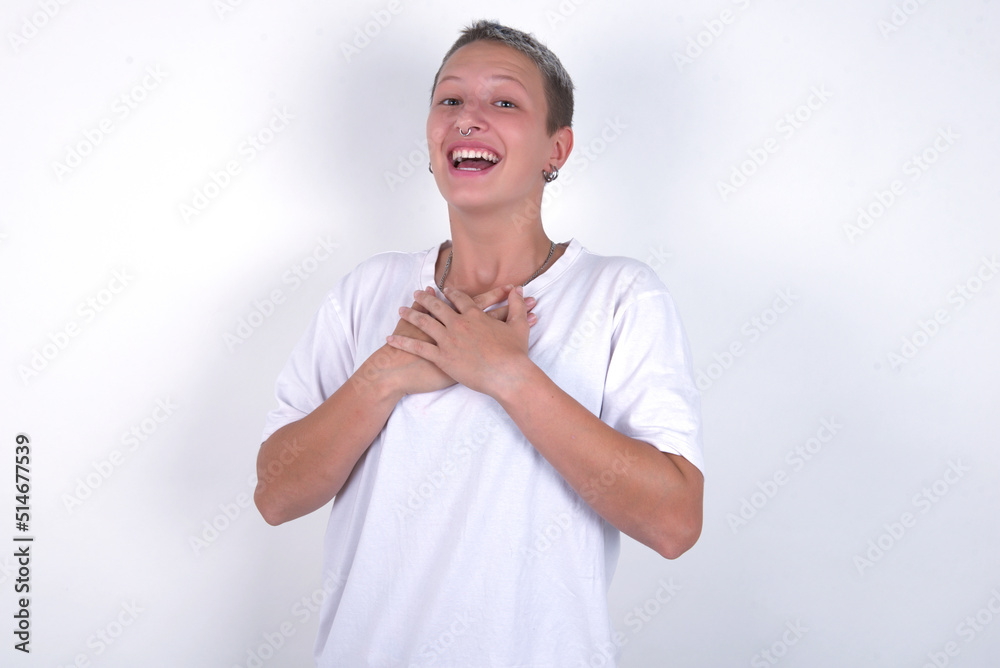 Happy smiling young woman with short hair wearing white t-shirt over white background has hands on chest near heart. Human emotions, real feelings and facial expression concept. Happy smiling young woman with short hair wearing white t-shirt over white background has hands on chest near heart. Human emotions, real feelings and facial expression concept.