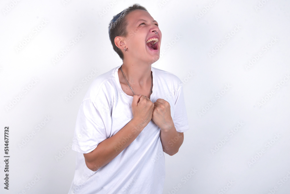 young woman with short hair wearing white t-shirt over white background excited and glad to achieve victory, clenches fists, screams in excitement with closed eyes,successful person. young woman with short hair wearing white t-shirt over white background excited and glad to achieve victory, clenches fists, screams in excitement with closed eyes,successful person.