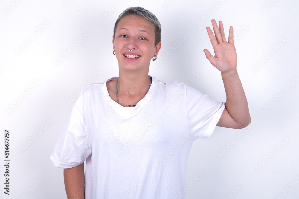 young woman with short hair wearing white t-shirt over white background waiving saying hello or goodbye happy and smiling, friendly welcome gesture.