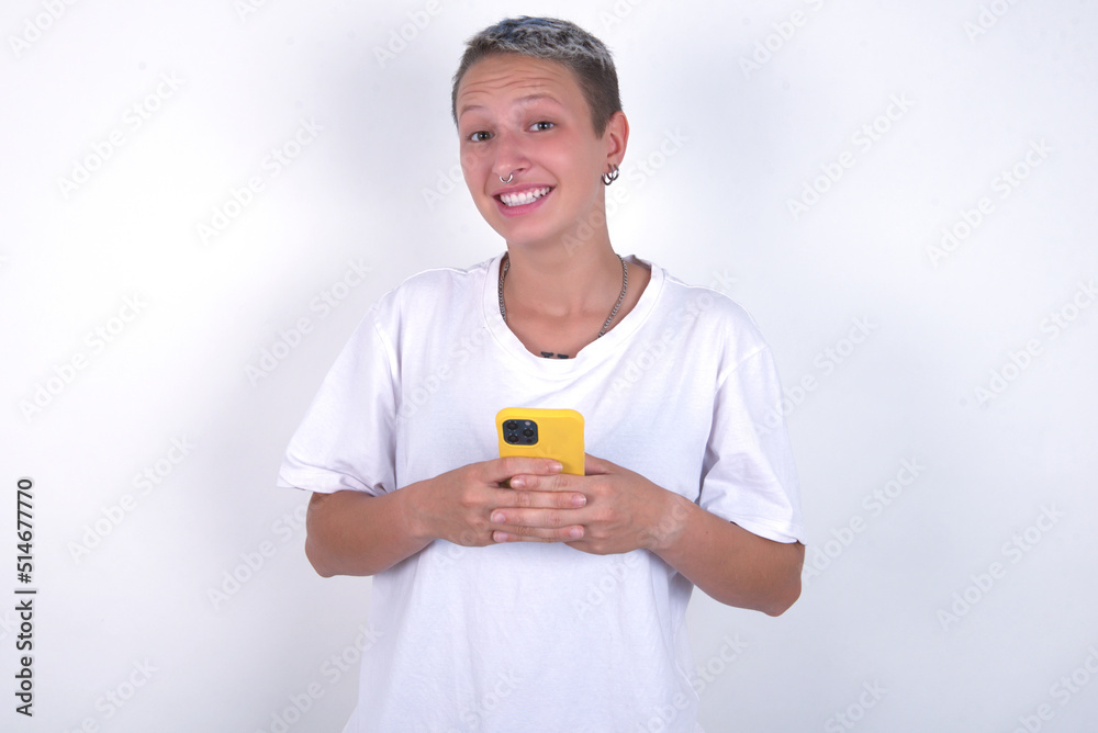 Smiling young woman with short hair wearing white t-shirt over white background friendly and happily holding mobile phone taking selfie in mirror. Smiling young woman with short hair wearing white t-shirt over white background friendly and happily holding mobile phone taking selfie in mirror.