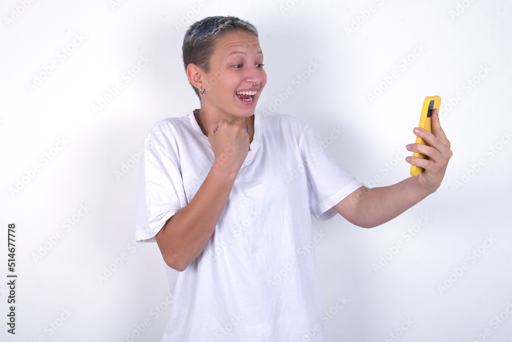Happy cheerful young woman with short hair wearing white t-shirt over white background receiving good news via e-mail and celebrating success while standing and looking at mobile phone. Happy cheerful young woman with short hair wearing white t-shirt over white background receiving good news via e-mail and celebrating success while standing and looking at mobile phone.