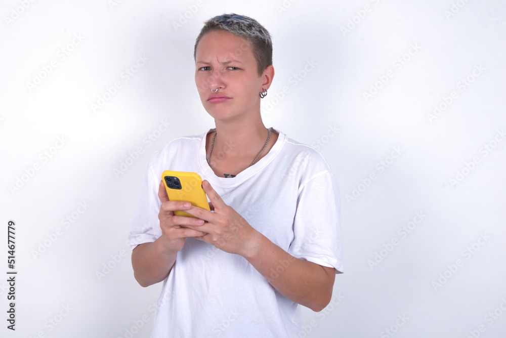 Portrait of a confused young woman with short hair wearing white t-shirt over white background holding mobile phone and shrugging shoulders and frowning face. Portrait of a confused young woman with short hair wearing white t-shirt over white background holding mobile phone and shrugging shoulders and frowning face.