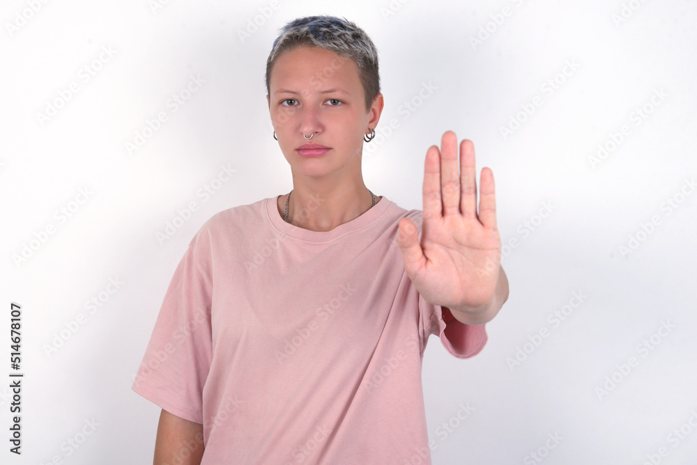 young woman with short hair wearing pink t-shirt over white background shows stop sign prohibition symbol keeps palm forward to camera with strict expression