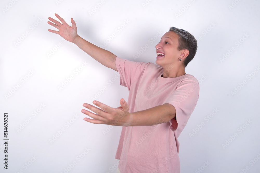 Funny astonished young woman with short hair wearing pink t-shirt over white background look empty space with arms opened ready to catch something. Funny astonished young woman with short hair wearing pink t-shirt over white background look empty space with arms opened ready to catch something.