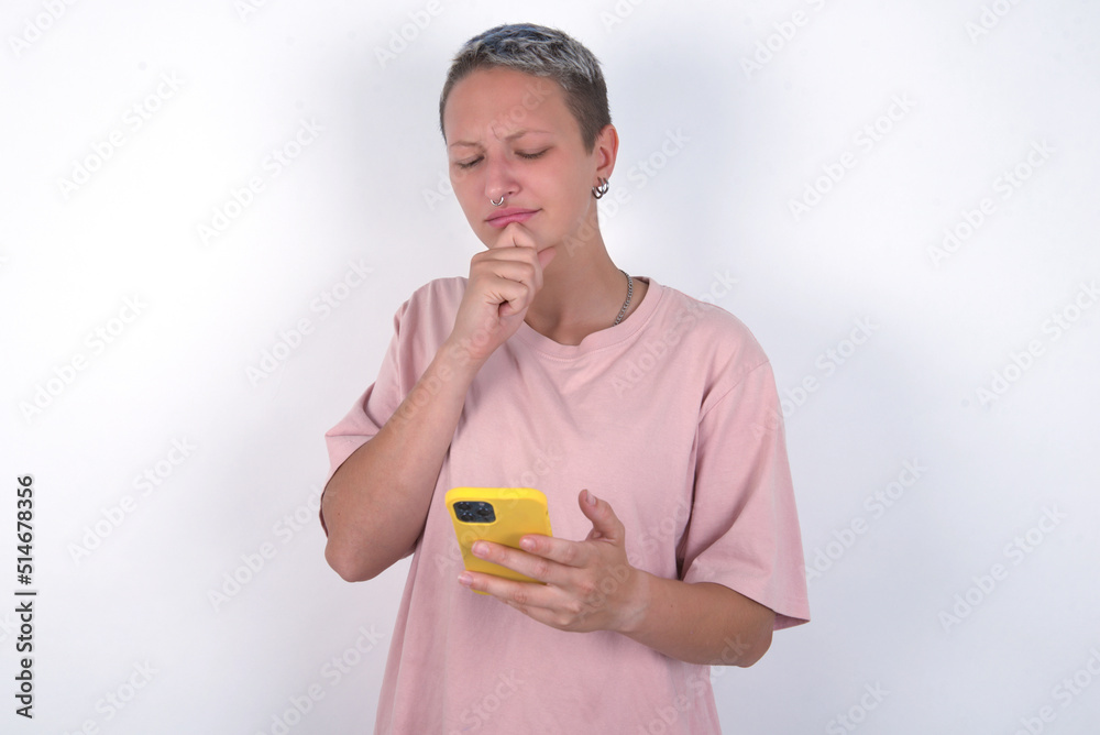 Thoughtful happy young woman with short hair wearing pink t-shirt over white background hold look phone, copyspace Thoughtful happy young woman with short hair wearing pink t-shirt over white background hold look phone, copyspace