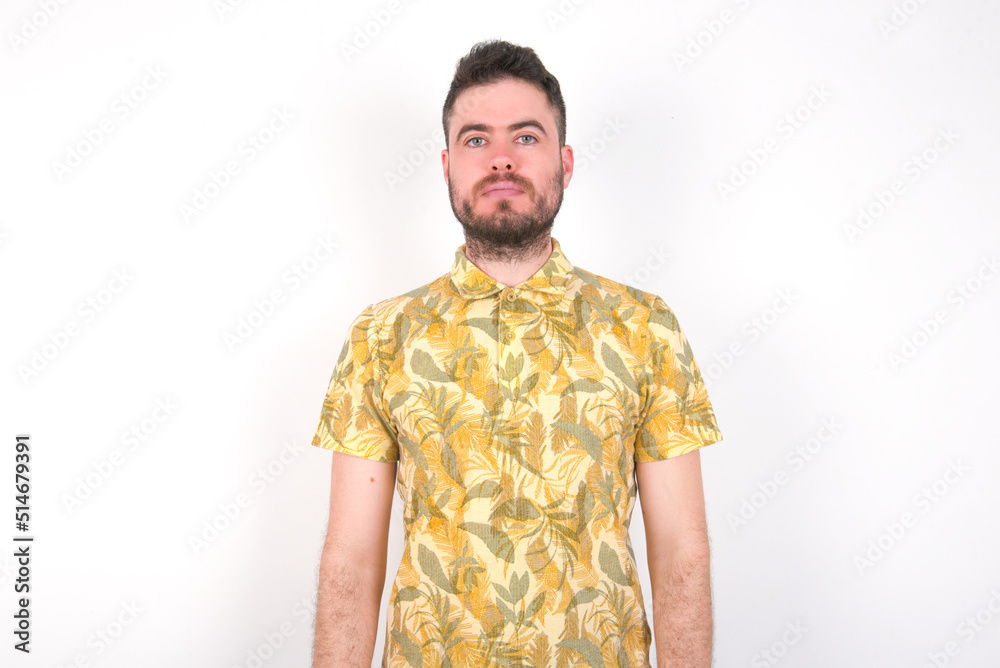Joyful Young caucasian man wearing Hawaiian t-shirt over white background  looking to the camera, thinking about something. Both arms down, neutral facial expression.