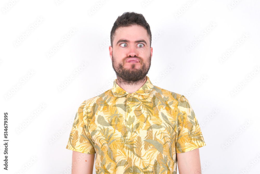 Young caucasian man wearing Hawaiian t-shirt over white background crosses eyes, puts lips, makes grimace with awkward expression has fun alone, plays fool. Young caucasian man wearing Hawaiian t-shirt over white background crosses eyes, puts lips, makes grimace with awkward expression has fun alone, plays fool.