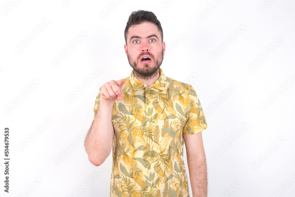 Fototapeta premium Shocked Young caucasian man wearing Hawaiian t-shirt over white background points front with index finger at camera and. Surprise and advertisement concept.