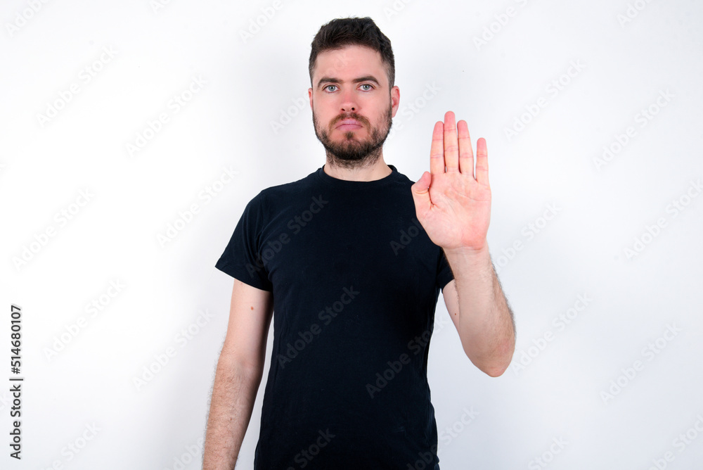 young caucasian bearded man wearing black t-shirt standing over white wall shows stop sign prohibition symbol keeps palm forward to camera with strict expression young caucasian bearded man wearing black t-shirt standing over white wall shows stop sign prohibition symbol keeps palm forward to camera with strict expression