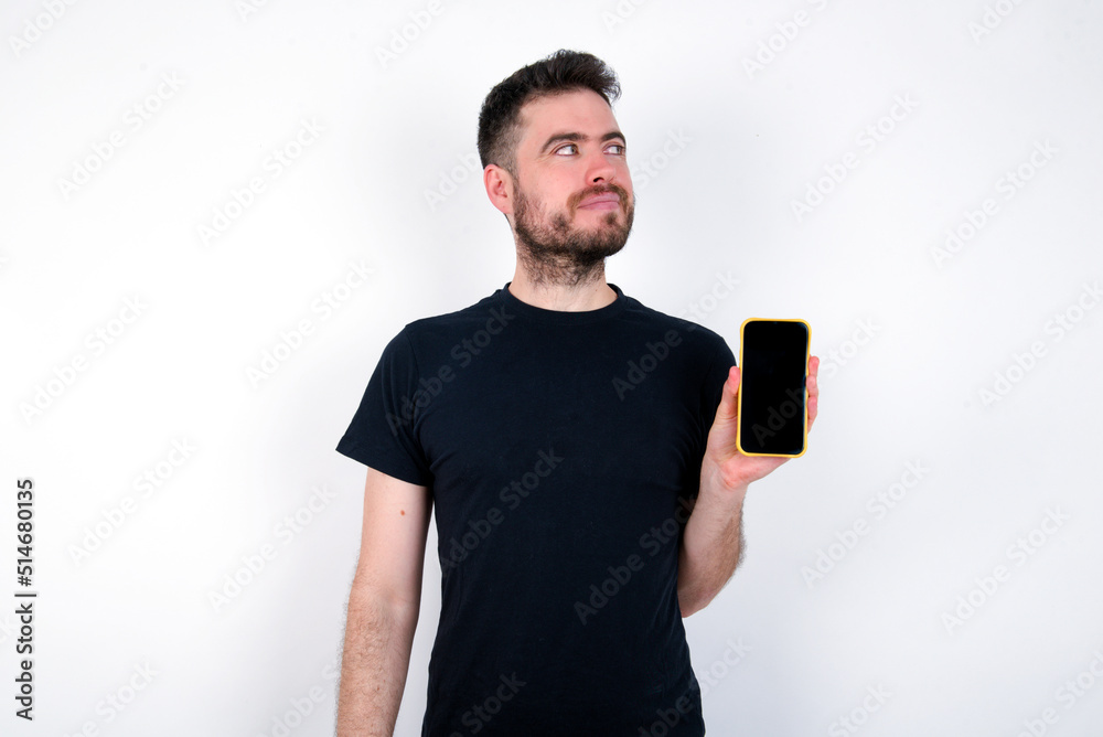 young caucasian bearded man wearing black t-shirt standing over white wall holds new mobile phone and looks mysterious aside shows blank display of modern cellular young caucasian bearded man wearing black t-shirt standing over white wall holds new mobile phone and looks mysterious aside shows blank display of modern cellular