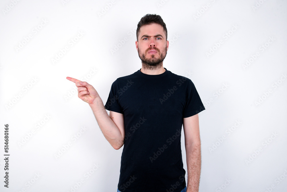 Serious young caucasian bearded man wearing black t-shirt standing over white wall smirks face points away on copy space shows something unpleasant. Look at this advertisement. Big price concept Serious young caucasian bearded man wearing black t-shirt standing over white wall smirks face points away on copy space shows something unpleasant. Look at this advertisement. Big price concept