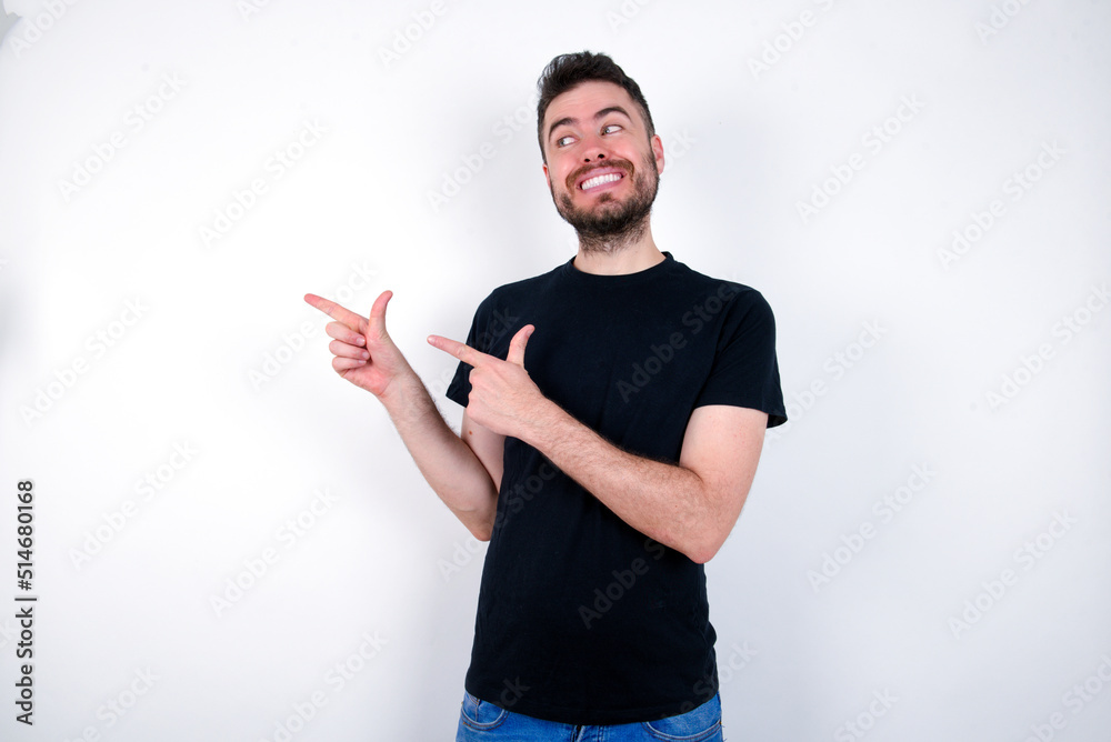 Optimistic young caucasian bearded man wearing black t-shirt standing over white wall points with both hands and looking at empty space. Optimistic young caucasian bearded man wearing black t-shirt standing over white wall points with both hands and looking at empty space.