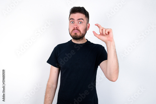 young caucasian bearded man wearing black t-shirt standing over white wall purses lip and gestures with hand, shows something very little.