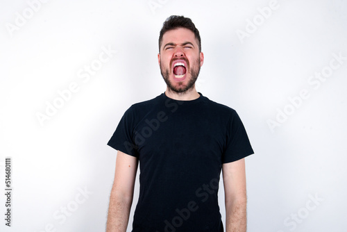 young caucasian bearded man wearing black t-shirt standing over white wall yawns with opened mouth stands. Daily morning routine © Jihan