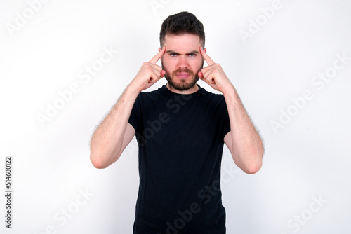 Serious concentrated young caucasian bearded man wearing black t-shirt keeps fingers on temples, tries to ease tension, gather with thoughts and remember important information for exam