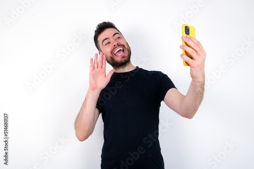 young caucasian bearded man wearing black t-shirt standing over white wall holds modern mobile phone and makes video call waves palm in hello gesture. People modern technology concept