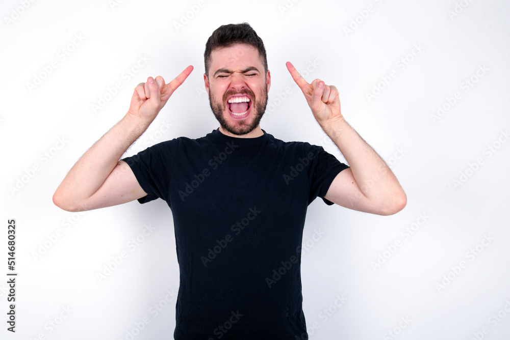 Fototapeta premium Photo of crazy young caucasian bearded man wearing black t-shirt standing over white wall screaming and pointing with fingers at hair closed eyes