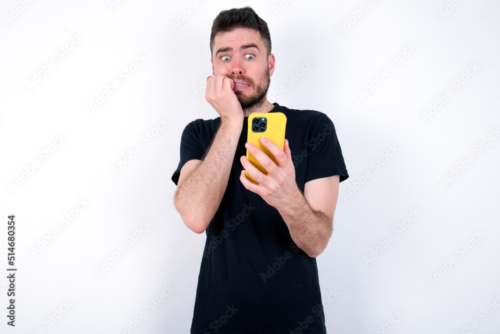 Portrait of pretty frightened young caucasian bearded man wearing black t-shirt standing over white wall chatting biting nails after reading some scary news on her smartphone.