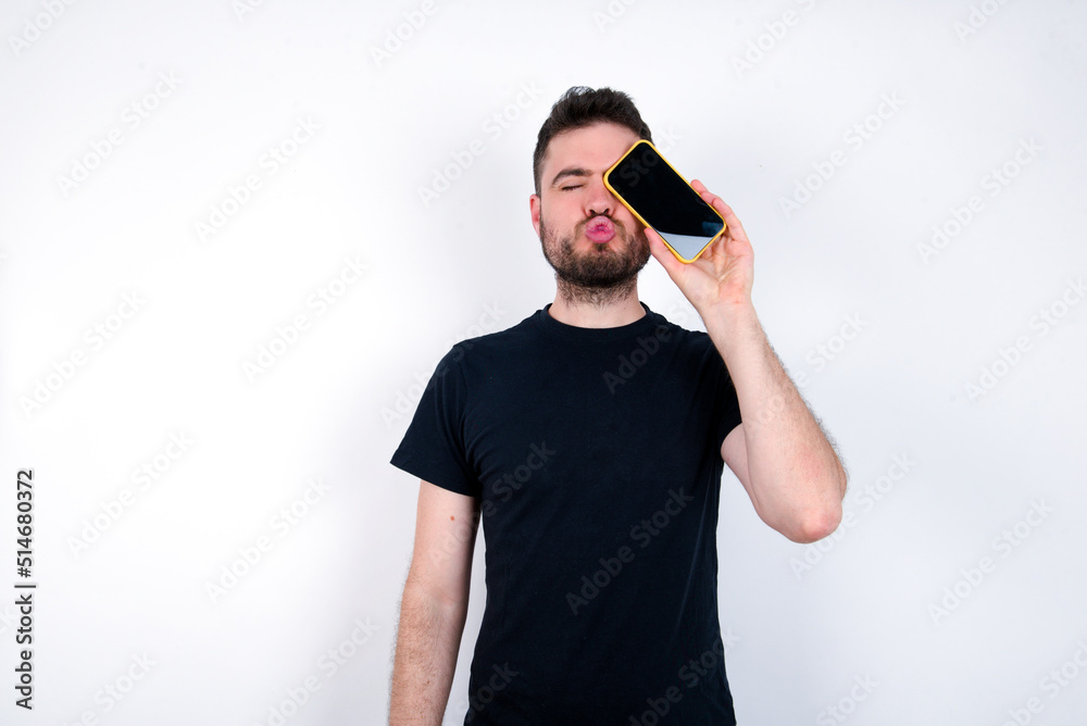 Adorable young caucasian bearded man wearing black t-shirt standing over white wall holding modern device covering eye with lips pouted Adorable young caucasian bearded man wearing black t-shirt standing over white wall holding modern device covering eye with lips pouted