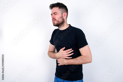 young caucasian bearded man wearing black t-shirt standing over white wall got stomachache
