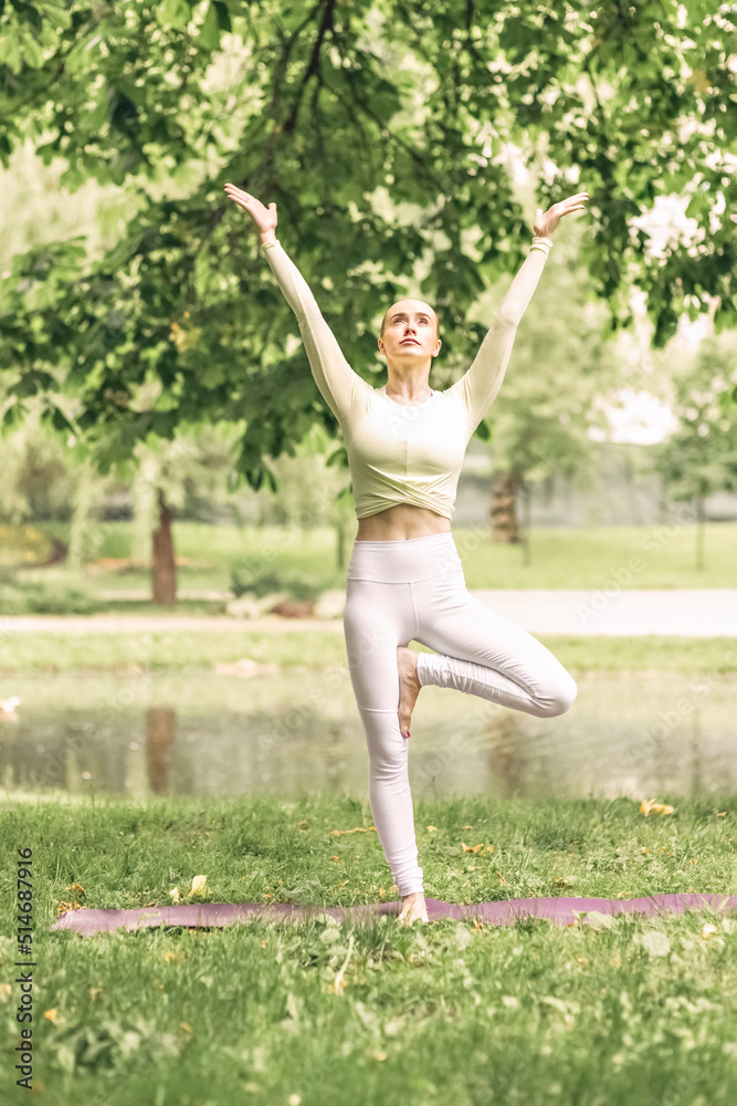 A young beautiful slender girl with long blonde hair does yoga in the summer in nature by the pond in the park.