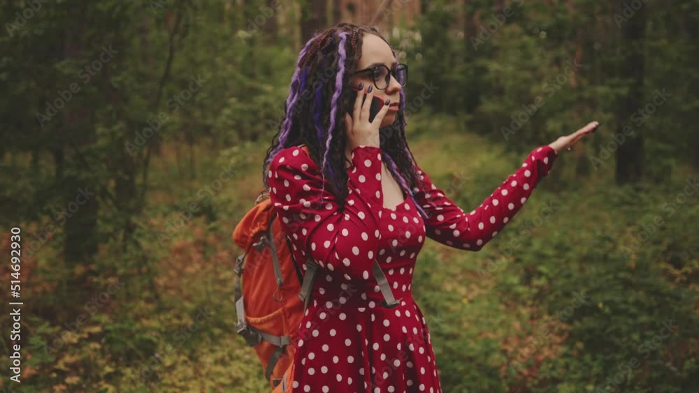 Lost young woman in glasses with backpack talking on mobile phone, standing in summer forest. Female tourist speaking on smartphone, stray in woods. Lost young woman in glasses with backpack talking on mobile phone, standing in summer forest. Female tourist speaking on smartphone, stray in woods.