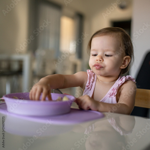 One child small caucasian toddler female baby eating at the table alone at home in room real people copy space early child development concept learning front view hold food healthy eating