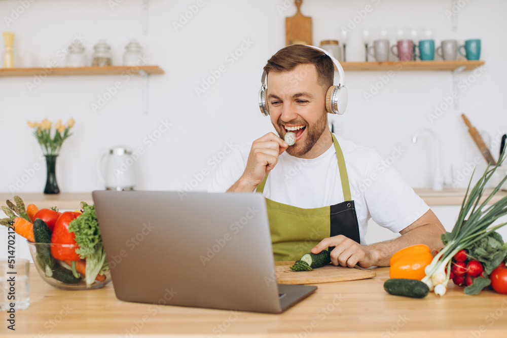 Happy man in headphones preparing salad in kitchen from fresh vegetables and looking at laptop. Happy man in headphones preparing salad in kitchen from fresh vegetables and looking at laptop.