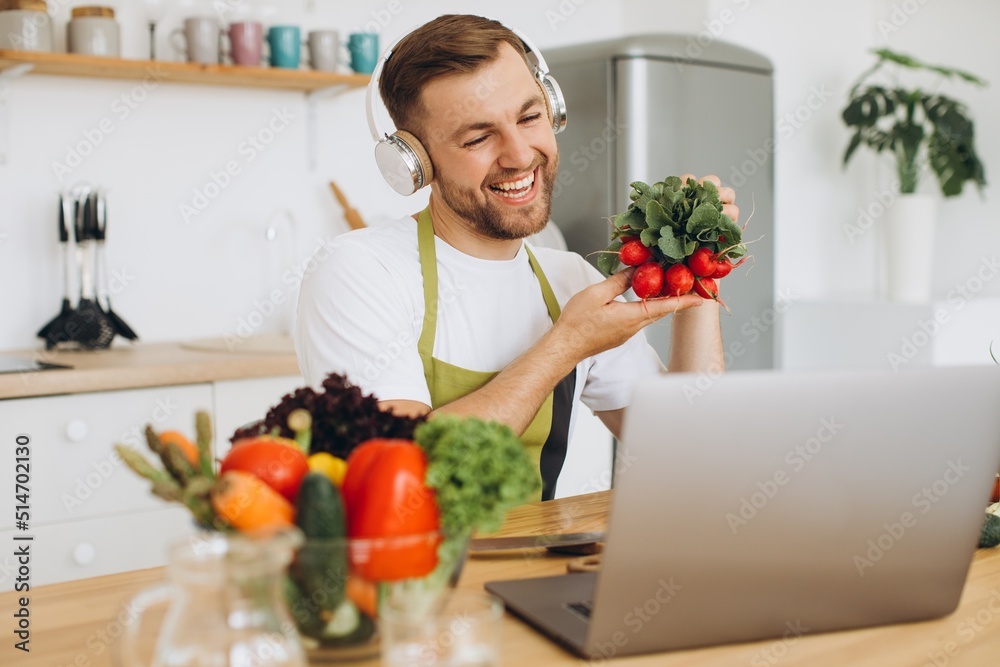 Happy man in headphones sitting at kitchen table and preparing salad, holding radish and showing it to laptop. Happy man in headphones sitting at kitchen table and preparing salad, holding radish and showing it to laptop.