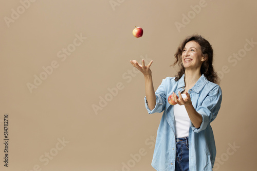 Happy smiling focused curly beautiful female in jeans casual shirt juggling with apple posing isolated on over beige pastel background. Healthy food. Natural eco-friendly products concept. Copy space