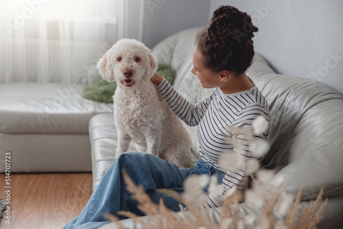 Young mixed-race woman owner sitting on couch at cozy house and pats her poodle dog