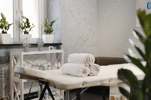 Rolled white towels on massage table in empty salon. Interior of aesthetic clinic, nobody. Modern cabinet of physiotherapist. Empty room with plants of beautician in wellness center. Copy space photo