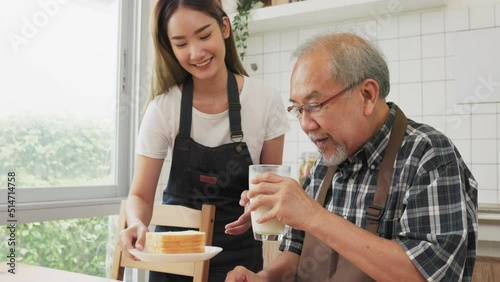 Asian lovely family, young daughter prepare breakfast for older father. Attractive female wear apron bake bread serve with milk to senior elderly dad sitting on eating table in kitchen at house.