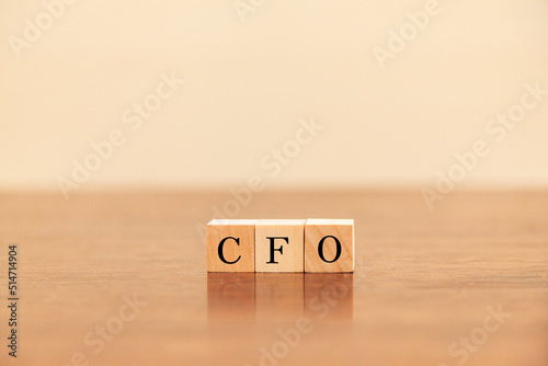 CFOの文字。最高財務責任者。Chief Financial Officer。3つの木製ブロックに書かれている。黒い文字。木製テーブルと白い壁紙の背景。
