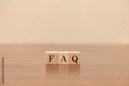 FAQの文字。よくある質問。Frequently Asked Questions。3つの木製ブロックに書かれている。黒い文字。木製テーブルと白い壁紙の背景。